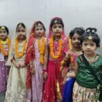 Six young children in bright traditional indian outfits pose together against a gray wall each wearing marigold garlands for a festive event